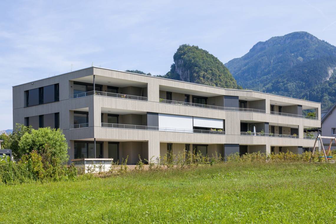 Das dreigeschossige Wohngebäude verbindet eine lasierte Holzfassade mit zurückversetzten Terrassen und großen Fensterflächen. Rasen, junge Pflanzungen und die umliegende Berglandschaft rahmen die Anlage ein. Wohnanlage Roseggerstraße in Hohenems