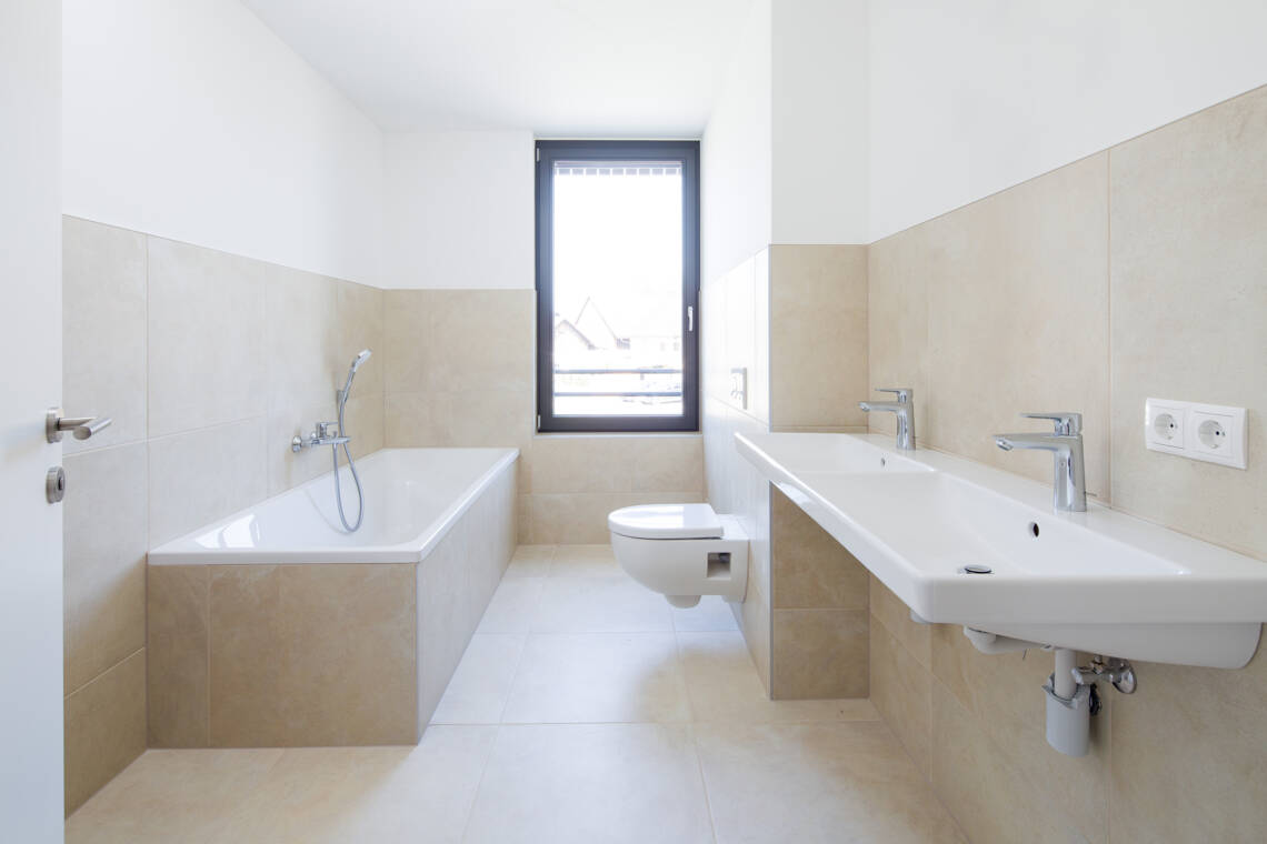 Das fertiggestellte Badezimmer verfügt über eine Badewanne mit Handbrause, einen breiten Doppelwaschtisch, ein wandhängendes WC und ein Fenster. Großformatiges Feinsteinzeug in warmem Beigeton prägt Boden und Wandflächen Badezimmer in Wohnung A7 mit Badewanne