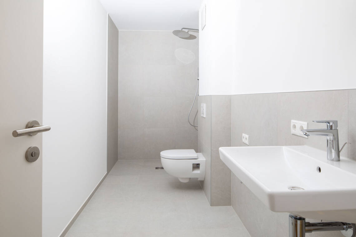 Das Badezimmer ist mit hellen Fliesen, wandhängendem WC, Waschbecken und einer bodengleichen Dusche mit Regenduschkopf ausgestattet. Badezimmer mit bodengleicher Dusche